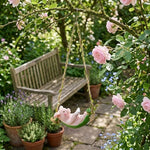 A pink ceramic bird swing hanging from a tree branch in a lush garden, with a wooden bench and potted plants in the background.