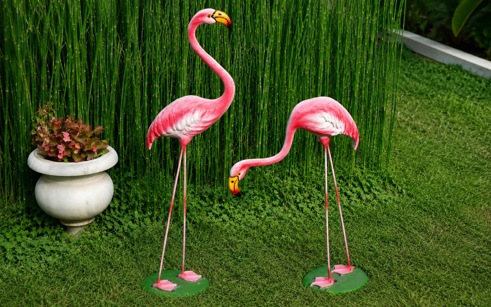 Two pink flamingo garden statues standing on green grass in front of a tall green hedge, with a white planter pot to the left.