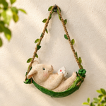 A hanging wall decor piece featuring a cream-colored rabbit curled up inside a green leaf-shaped hammock, suspended by a rope with small green leaves, against a textured off-white wall.