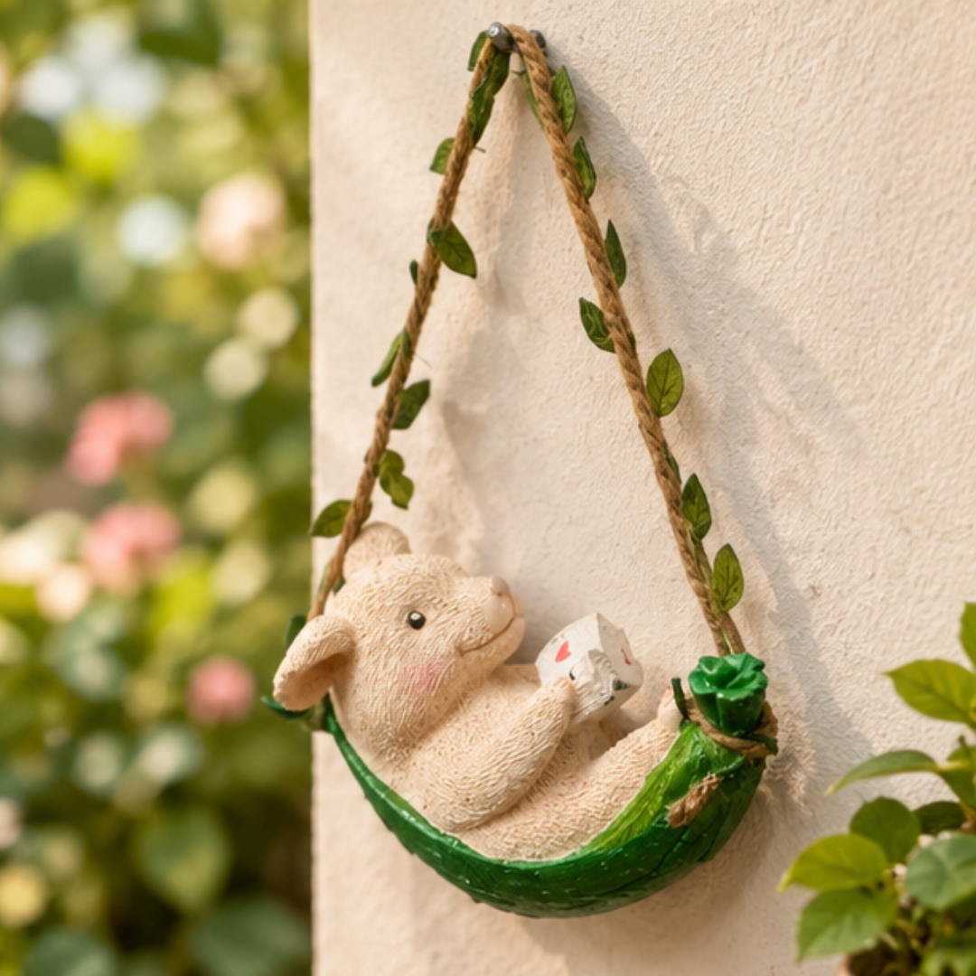 A decorative hanging ornament featuring a cream-colored bunny sitting in a green leafy hammock suspended from a rope, displayed against a textured beige wall with blurred green foliage in the background.