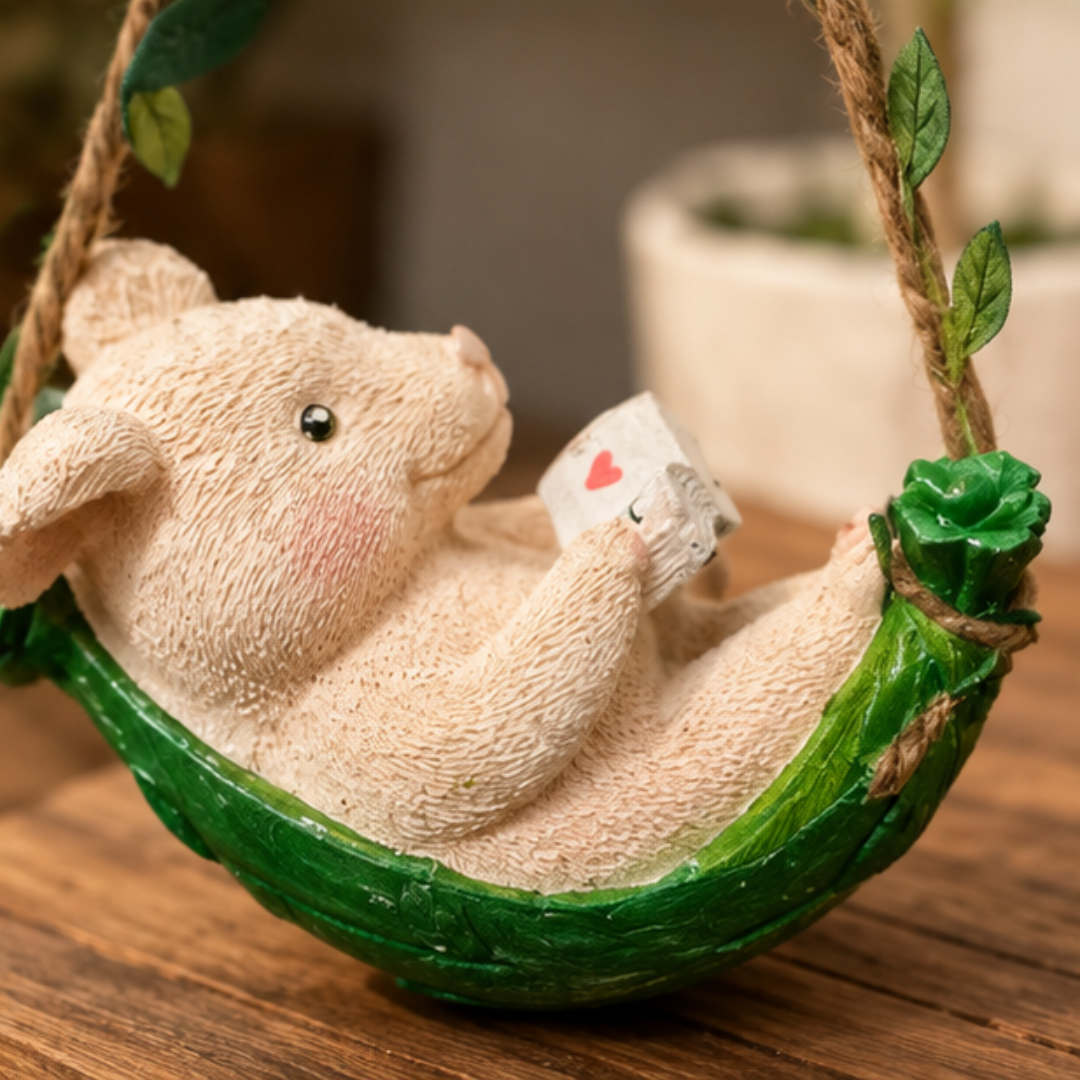 A small beige animal figurine sitting in a green leafy hammock swing, holding a tiny card, photographed on a wooden surface with a softly blurred indoor background.