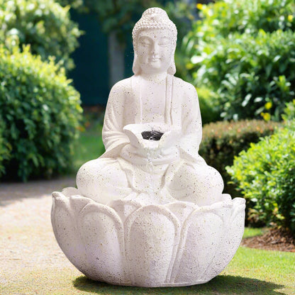 A white stone-looking Buddha water fountain with LED lighting placed on a wooden surface with greenery in the background.