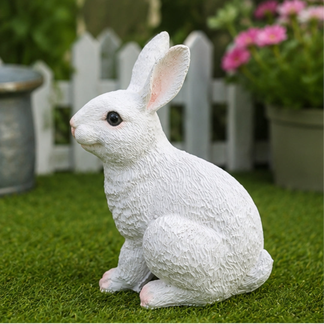 A white textured rabbit garden statue sitting on green grass in front of a white picket fence and potted flowers.