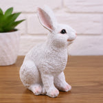 A white textured rabbit figurine sitting on a wooden tabletop with a white brick wall and a potted plant in the background.