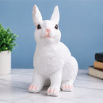 A white rabbit figurine with textured fur, pink nose and paw pads, sitting on a marble surface against a blue background with a small potted plant and books in the background.